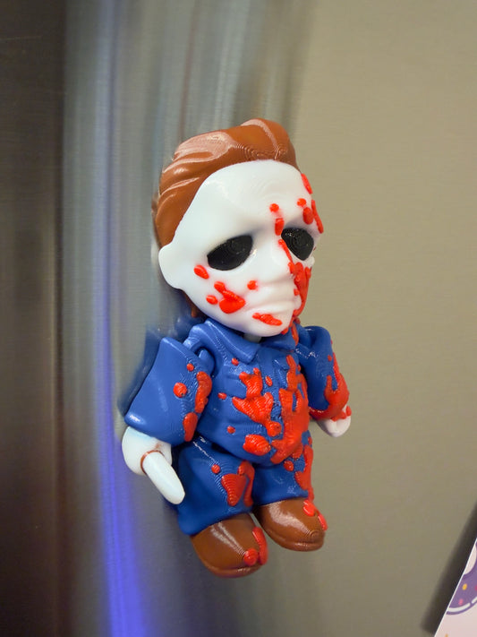 A small figurine or doll posed on its side, with a white face, brown hair, a blue outfit, and orange splatter details, photographed against a plain indoor background.