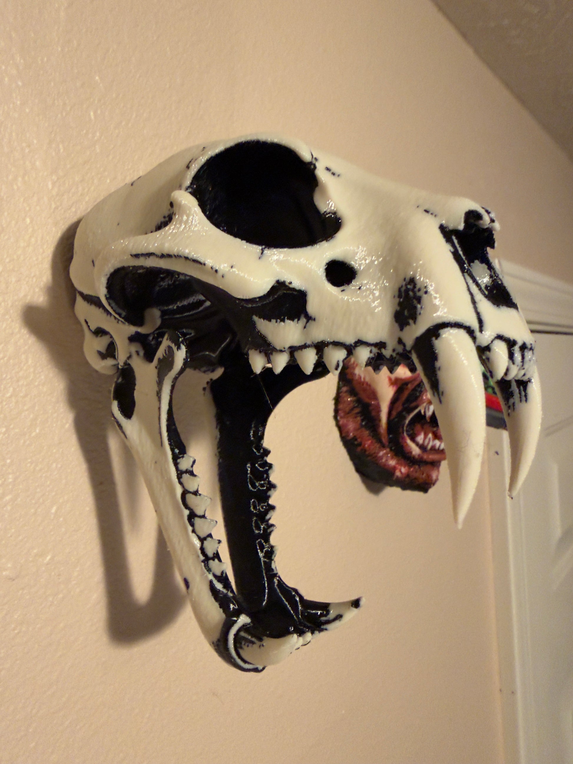 A 3D-printed animal skull replica, likely a wolf or coyote skull, featuring black and white painted details and large curved teeth. A small printed graphic of a human eye is visible inside the mouth.
