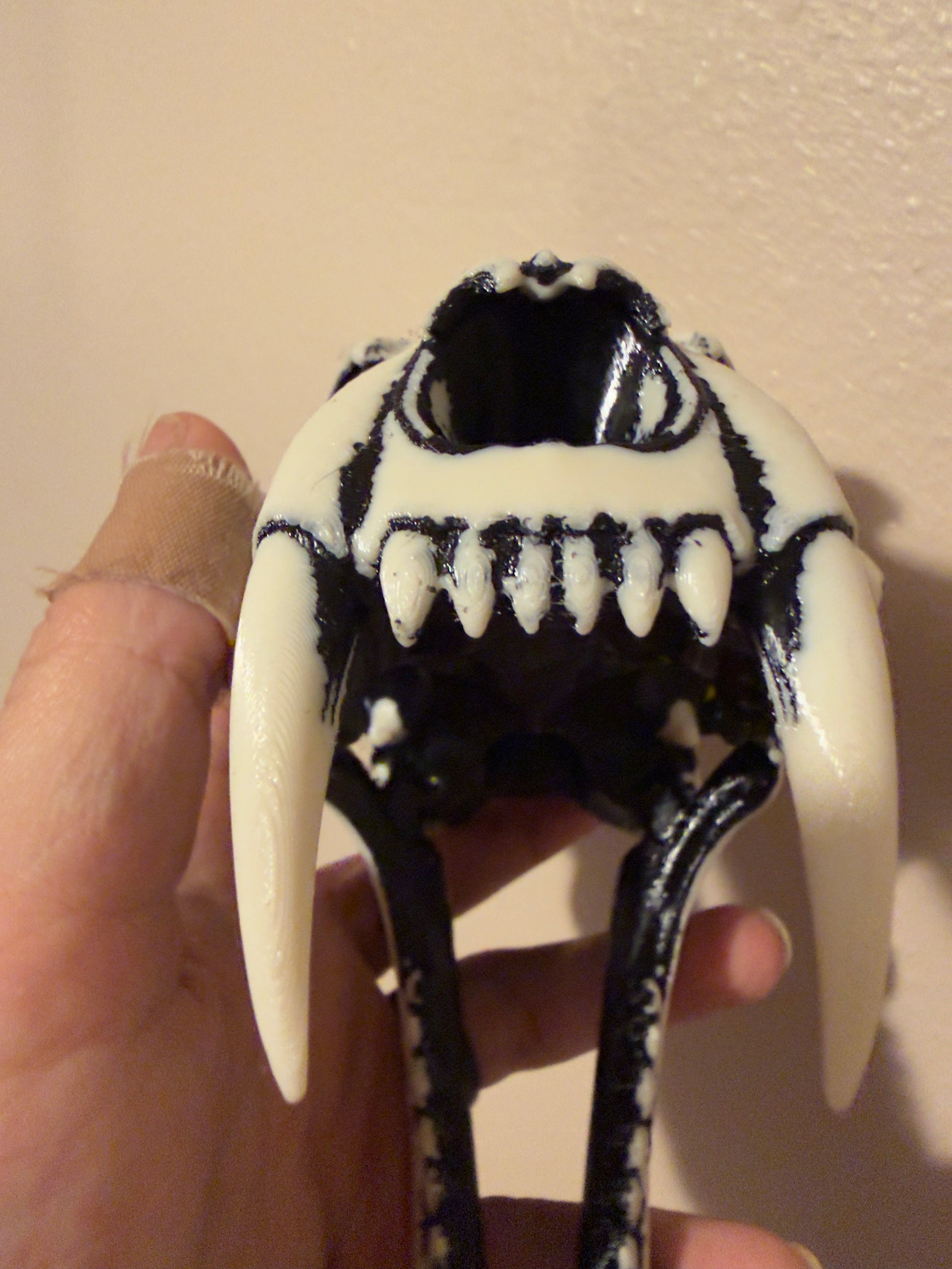 A close-up of a hand holding a black and white skull-shaped object with long curved fangs and sharp teeth, against a plain beige wall.