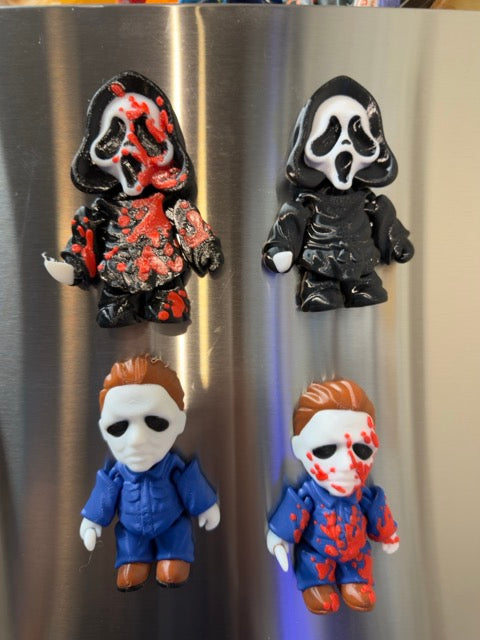 Four small horror character magnets or figurines displayed on a stainless steel refrigerator door. The set includes two Ghostface-style figures and two Michael Myers-style figures, with black, white, red, and blue color schemes.