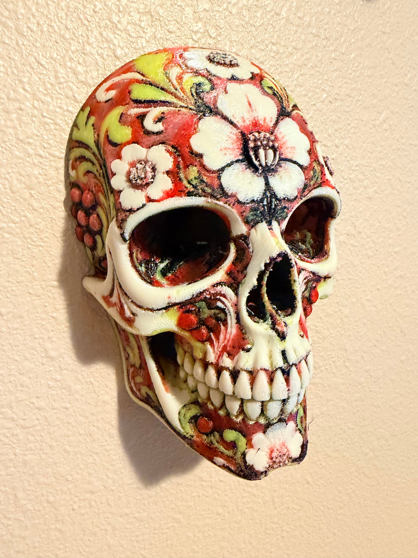 A decorative ceramic sugar skull figurine featuring a hand-painted floral pattern in red, white, green, and black, displayed on a textured beige surface.