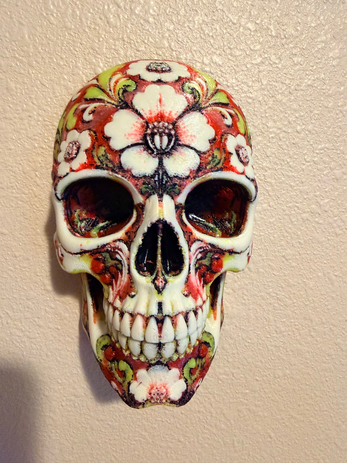 A decorative skull wall hanging with a sugar skull design, featuring white base with red, green, and black painted floral and scroll patterns, mounted on a textured beige wall.