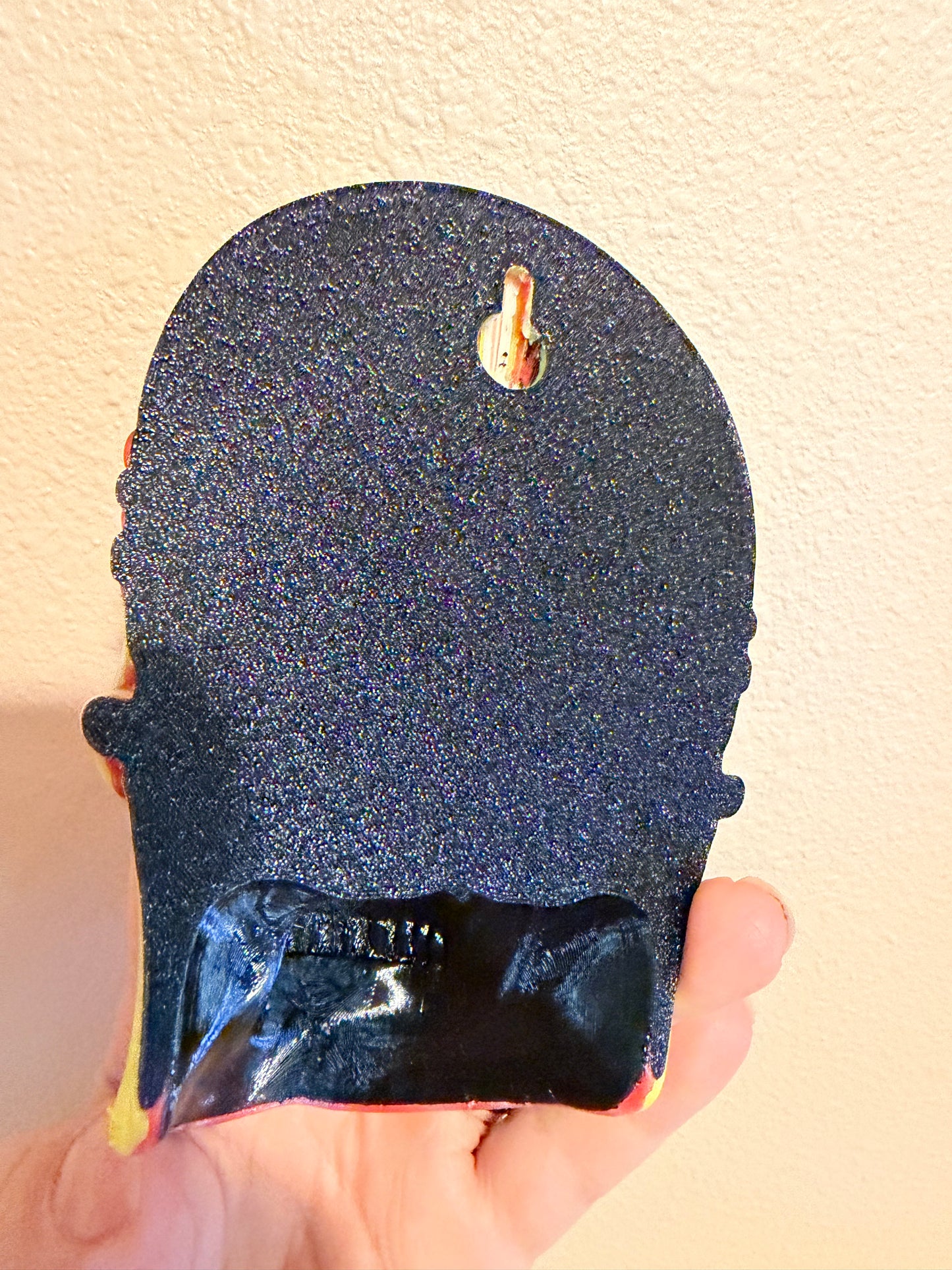 A hand holding a black glittery shoe-shaped object, likely a shoe tree or decorative shoe holder, against a textured beige wall.