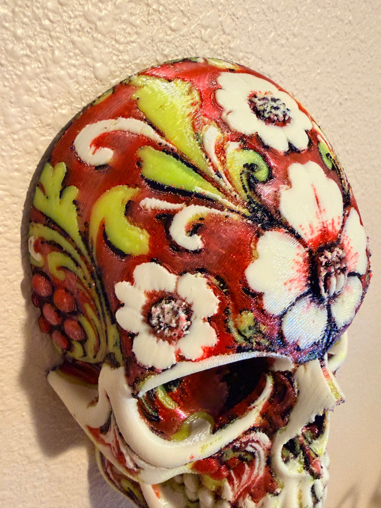 A decorative skull sculpture featuring a hand-painted Mexican Day of the Dead style design with white flowers, green leaves, and red marbled patterns on a textured beige wall.