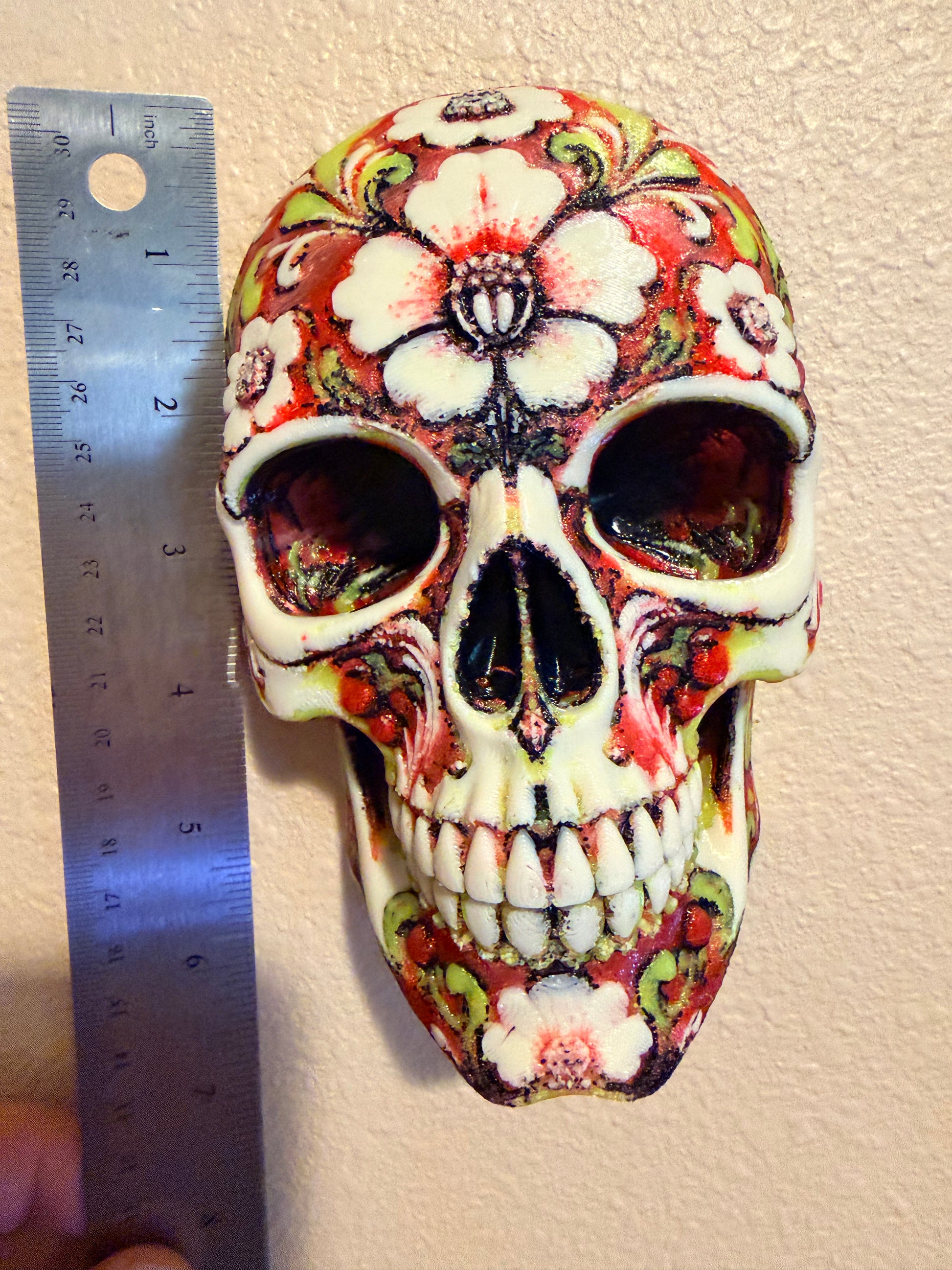 A decorative skull with a floral sugar skull pattern in red, white, green, and black, shown next to a metal ruler for scale against a textured beige wall.